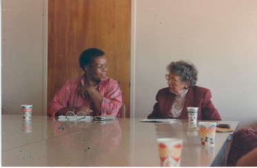 Thoko Ginindza and Hilda Kuper, 1984/85, from the Kuper family collection, courtesy of Mary Kuper. In this photo, Thoko is on the left, a Black woman with short black hair, in a red collared 3/4 length blouse with her left arm bent at the elbow, touching her necklace or chest. Her right hand and forearm are resting on the table in front of her. Her glasses, glass case, and a pad of paper are in front of her on the table. To her left, but on the viewer's right, is an older white woman with grey and white hair, Hilda Kuper, wearing a burgundy-colored jacket and a multi-colored high-necked blouse. She wears glasses and looks over at Thoko. She has a small disposable cup in front of her on the table. It looks to be a conference room of some kind. They are deep in conversation.
